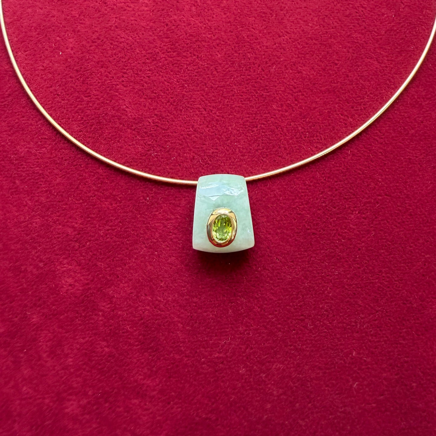 A delicate gold wire necklace with a pale green jade pendant featuring a gold bezel-set peridot, displayed on a textured dark red background.