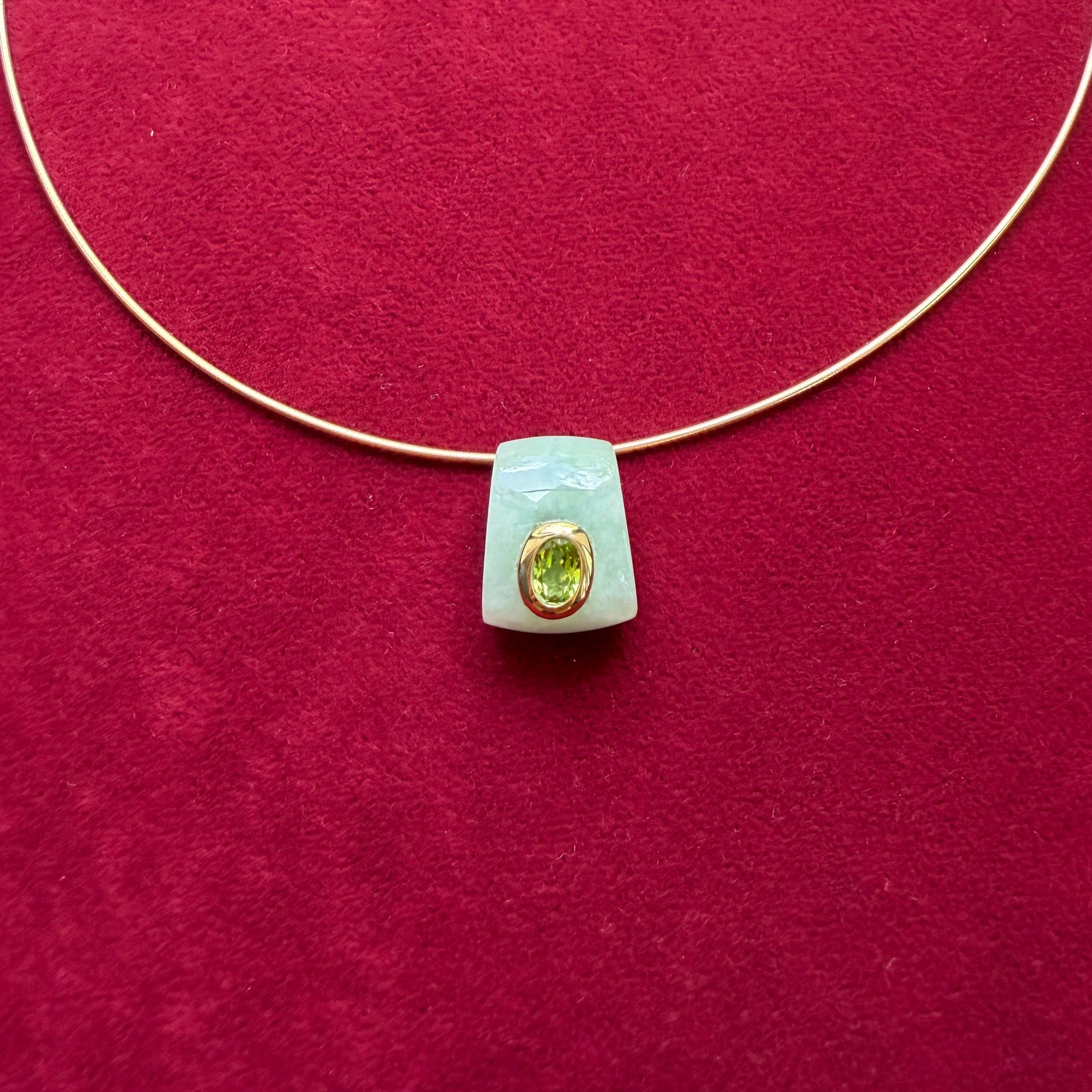 A delicate gold wire necklace with a pale green jade pendant featuring a gold bezel-set peridot, displayed on a textured dark red background.
