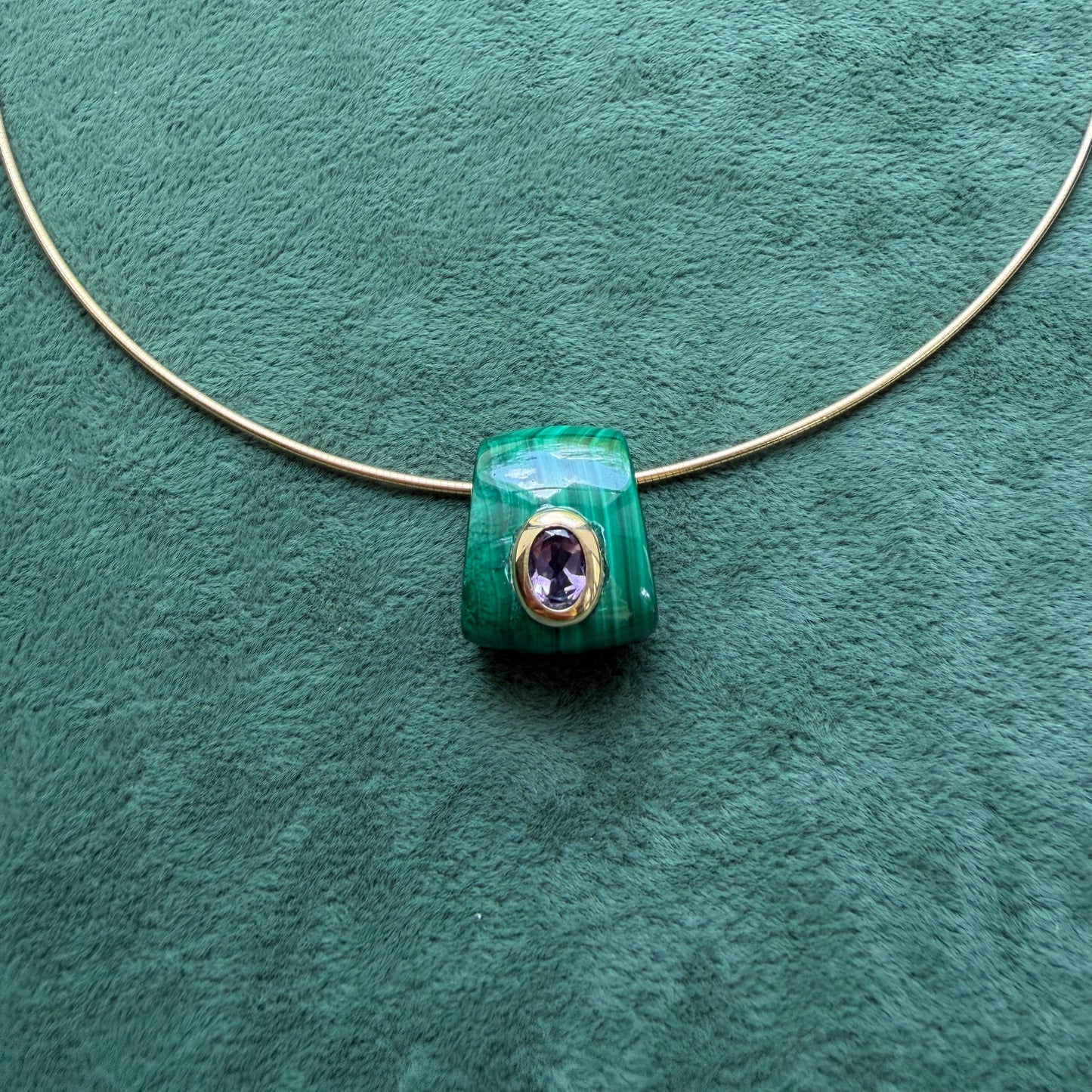A green malachite pendant featuring an amethyst gemstone, displayed on a textured green background.