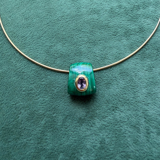 A green malachite pendant featuring an amethyst gemstone, displayed on a textured green background.
