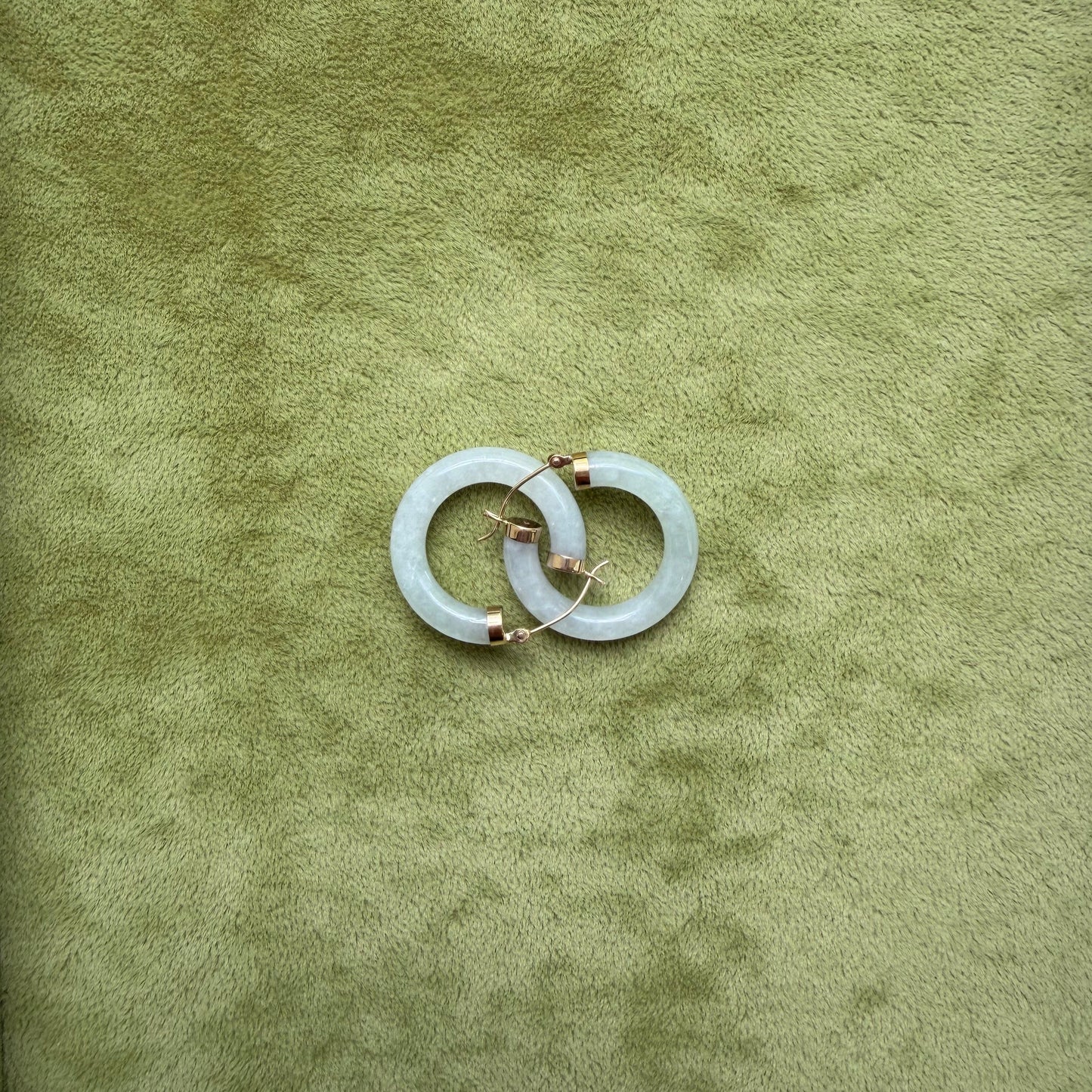 Two light green jade hoops with small 14k gold accents, arranged in an interlocking circular pattern on a textured olive green background.
