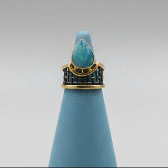 An teardrop shaped opal ring with a gold-toned band at the top, centered on a neutral gray studio background.