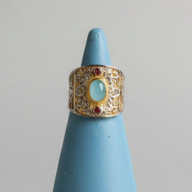 An ornate sterling silver ring with a central oval aquamarine stone and small red sapphires, displayed on a light blue cone stand against a neutral gray background.