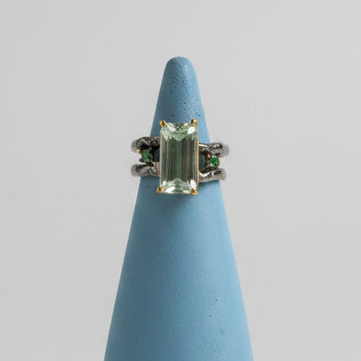 A silver ring with a central aquamarine emerald cut stone and smaller emerald side stones, displayed on a light blue cone-shaped stand against a neutral gray background.